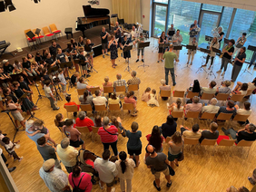 Ein kleines Orchester spielt in einem hellen Saal mit Holzboden vor einem sitzenden und stehenden Publikum.